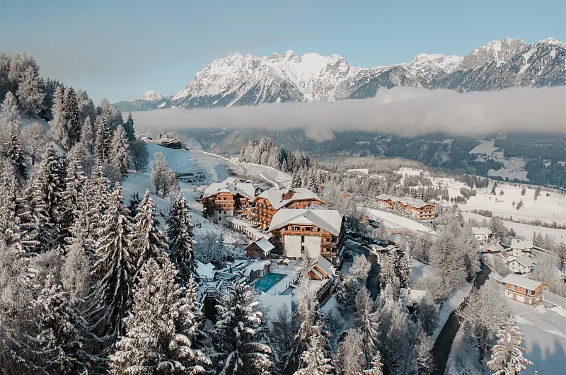 Drohnenaufnahme eines Hotels am verschneiten Waldhang mit Blick ins Tag