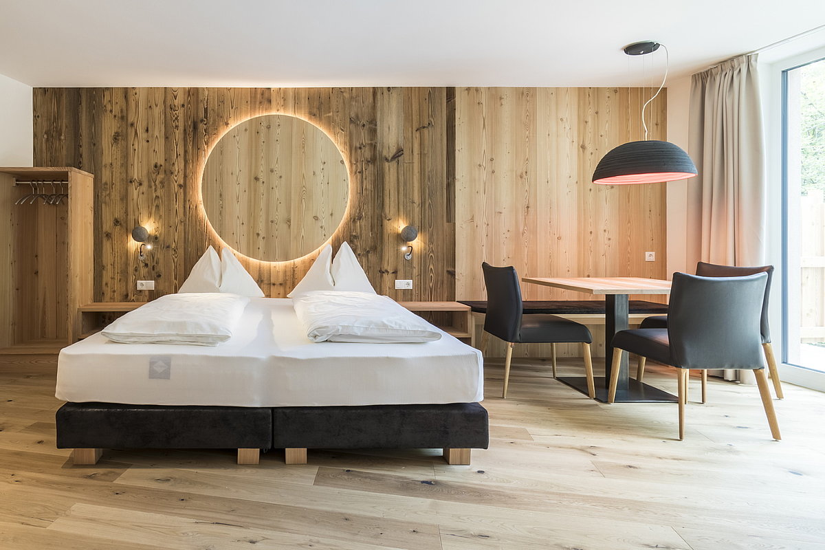 Auf dem Bild sieht man ein Hotelzimmer des Biohotel Steineggerhof. Man sieht viel Holz, z. B. an den Wänden. Die Einrichtung (Doppelbett) und Stühle mit Tisch sind sehr modern und stylisch gehalten.