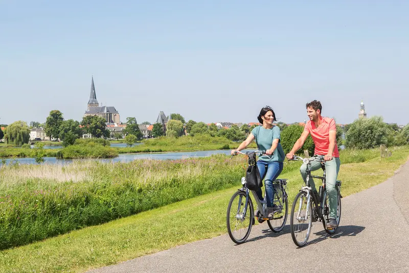 ein Mann und eine Frau radeln nebeneinander auf einem Radweg am Fluss entlang, im Hintergrund sind am gegenüberliegenden Flussufer eine Kirche und Häuser zu sehen