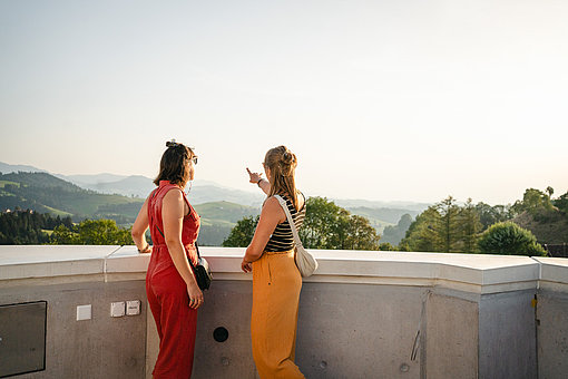 Zwei Frauen stehen an der Brüstung einer Dachterrasse und schauen in die Umgebung