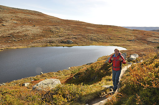 Ein Wanderer mit einer roten Jacke läuft in Norwegen an einem See entlang.