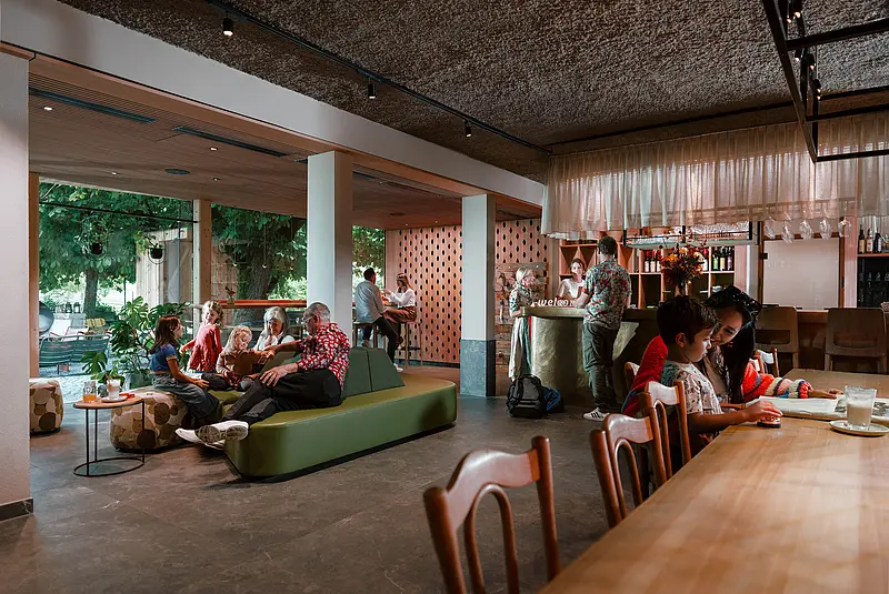 Hotellobby mit Tischen, Stühlen, Bänken, einer Bar und einem Sofa mit Menschen unterschiedlichen Alters