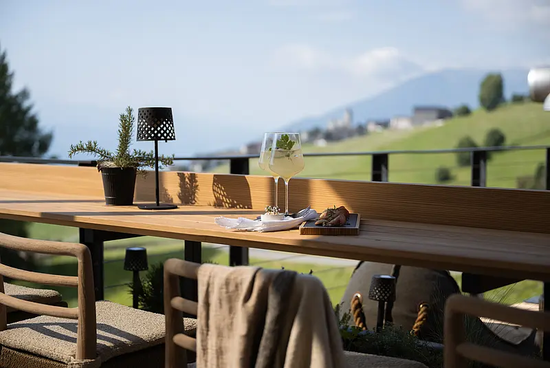 Auf einer sonnigen Hotelterrasse stehen ein langer Holztisch und gepolsterte Stühle. Auf dem Tisch stehen ein Getränk mit Kräutern und ein kleiner Snack, im Hintergrund sind grüne Wiesen und die unscharfe Südtiroler Berglandschaft zu sehen.