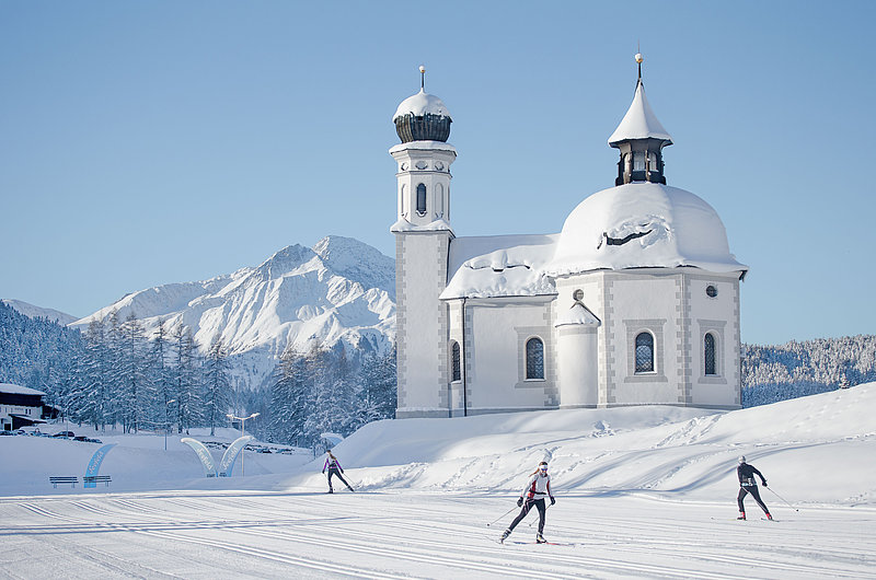 Eingeschneite Kirche in winterlicher Berglandschaft, im Vodergrund drei Langläufer