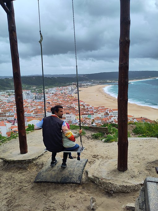 Ein Vater schaukelt mit seiner Tochter. Von der Schaukel aus hat man einen guten Blick über das Meer und eine Küstenstadt.
