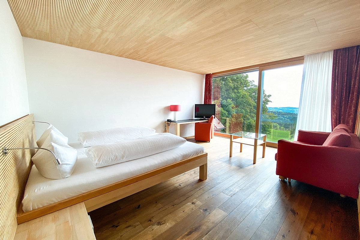 Ein Bett, ein Sessel und ein Schreibtisch mit Fernseher, in einem Zimmer mit Holzboden und -decke sowie Panorama-Ausblick