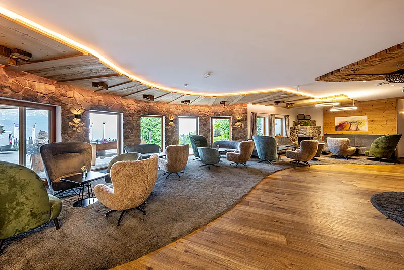 Plüschsessel stehen um kleine runde Couchtische auf einem Teppich in einem weitläufigen Raum mit großen Fenstern und Holzfußboden