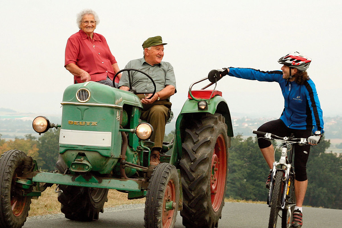 Ein älterer Mann und eine ältere Frau sitzen auf einem Traktor und nehmen einen Radfahrer ein Stück mit, der sich auf seinem Fahrrad während der Fahrt am Traktor festhält