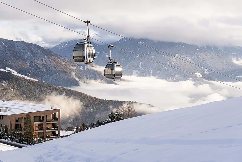 Zwei Gondeln einer Bergbahn schweben durch die Winterlandschaft, im Hintergrund ist das Hotel zu sehen
