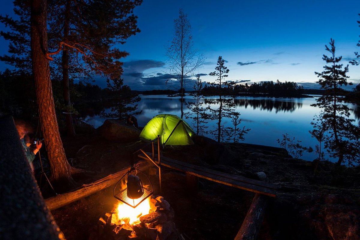 Ein grünes Zelt steht einsam inmitten von wilder Natur, im Vordergrund ist ein Lagerfeuer und im Hintergrund ein See zu sehen.