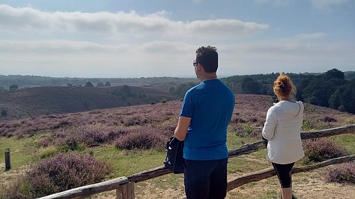 Ein Mann und eine Frau stehen auf einem Aussichtshügel und blicken über eine Heidelandschaft