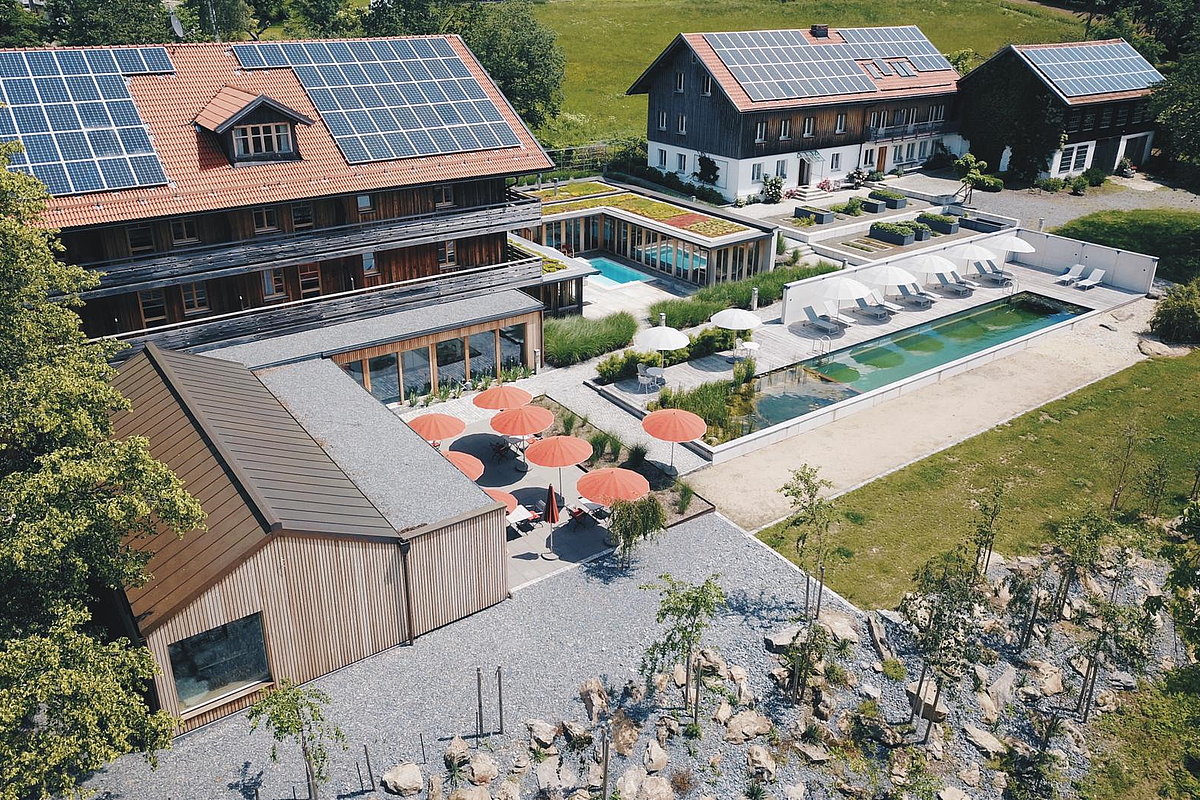 Gebäudekomplex bestehend aus vier mit Holz verkleideten Gebäuden mit Solaranlagen aus der Vogelperspektive, Kiesgarten, Sonnenschirme, Pool, Sonnenliegen