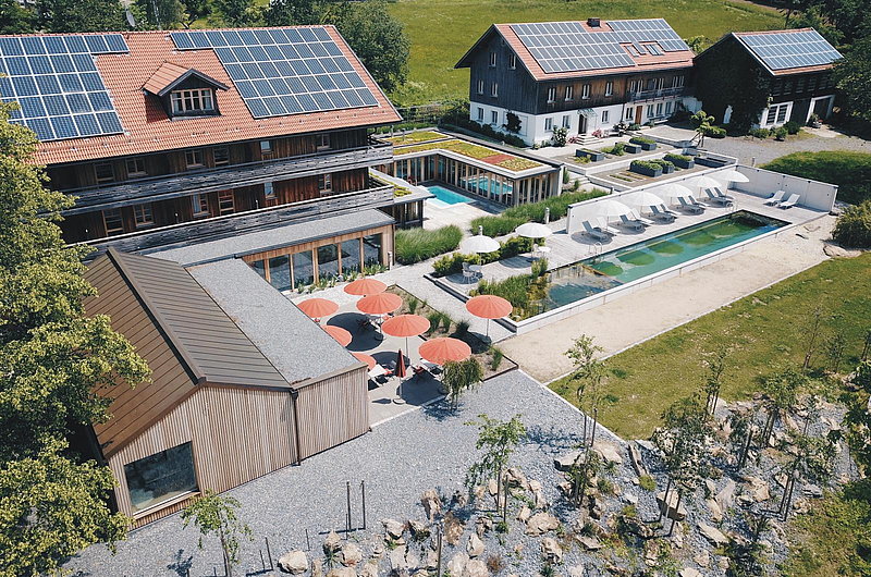 Gebäudekomplex bestehend aus vier mit Holz verkleideten Gebäuden mit Solaranlagen aus der Vogelperspektive, Kiesgarten, Sonnenschirme, Pool, Sonnenliegen
