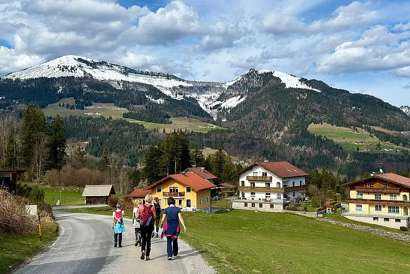 Fünf Frauen mit Tagesrucksäcken wandern auf einem Asphaltweg zwischen Häusern und Wiesen. Im Hintergrund bewaldete Berge mit Schneeresten auf den Gipfeln