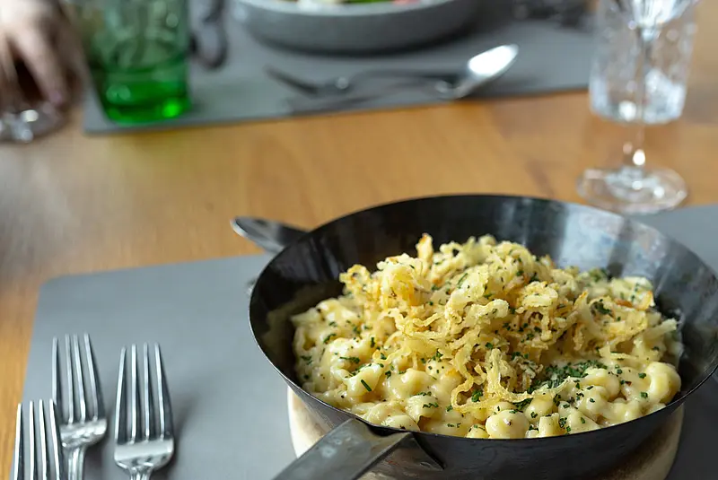 Eine Pfanne mit Käsenudeln steht auf deinem Holzbrett auf einem gedeckten Restauranttisch