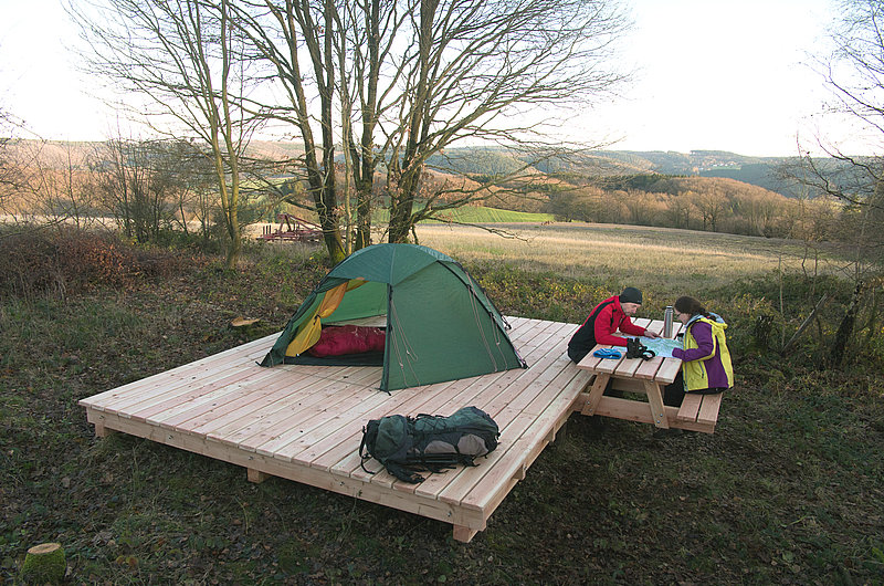 Zwei Menschen sitzen an einem Holz-Campingtisch neben einem bunten Zelt in einer herbstlichen Landschaft und blicken auf einer Landkarte