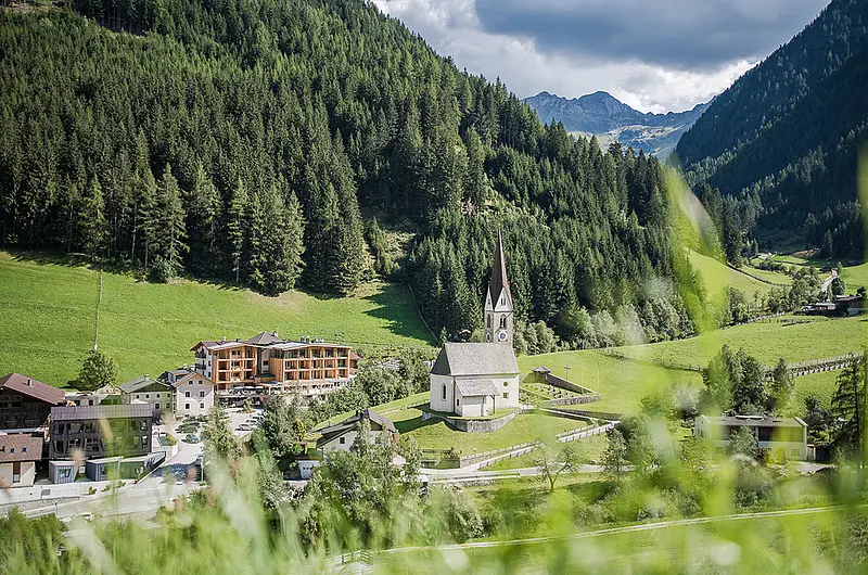 Luftaufnahme eines Hotels neben einer Dorfkirche umgeben von Wiesen, bewaldeten Berghängen und Hochgebirgsgipfeln