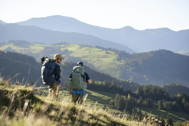 Zwei Männer wandern jeder mit einem Trekkingrucksack auf dem Rücken über eine Bergwiese ins Tal. Im Hintergrund bewaldete Hügel