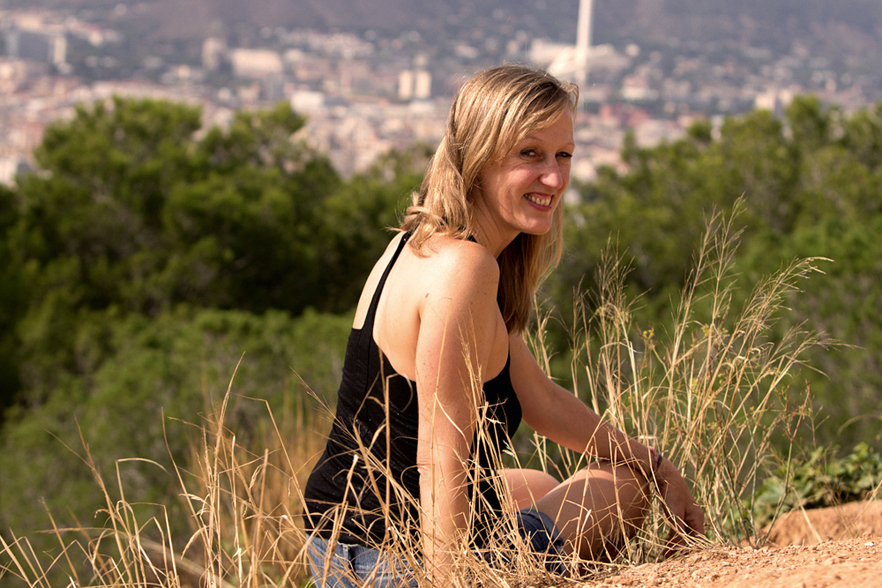 Eine junge Frau sitzt bei Sonnenschein im Gras und lächelt in die Kamera, im Hintergrund erstreckt sich Barcelona