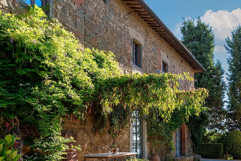 Blick in einen gepflasterten Hof mit begrünter Pergola, Sitzmöbeln und einem Steinhaus