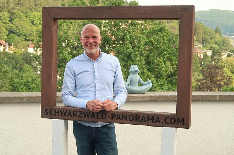 Ein Mann steht in einem großen Fotorahmen mit der Aufschrift "schwarzwald-panorama.com", hinter ihm befindet sich das Panorama des Schwarzwaldes