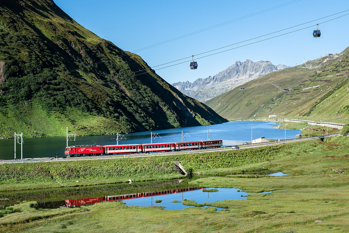 Ein Personenzug fährt in einem schweizer Alpental an einem See entlang.