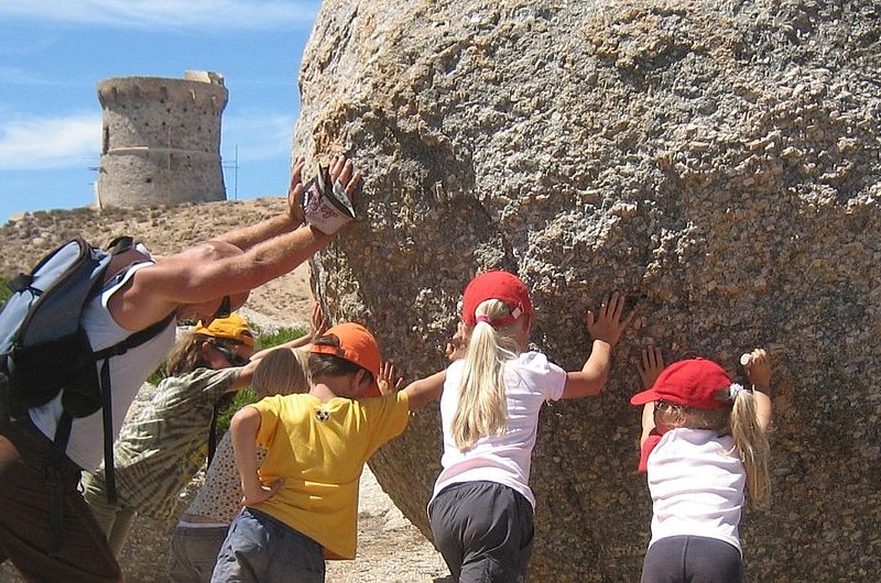 Eine erwachsene Person und drei Kinder schieben einen großen Stein.