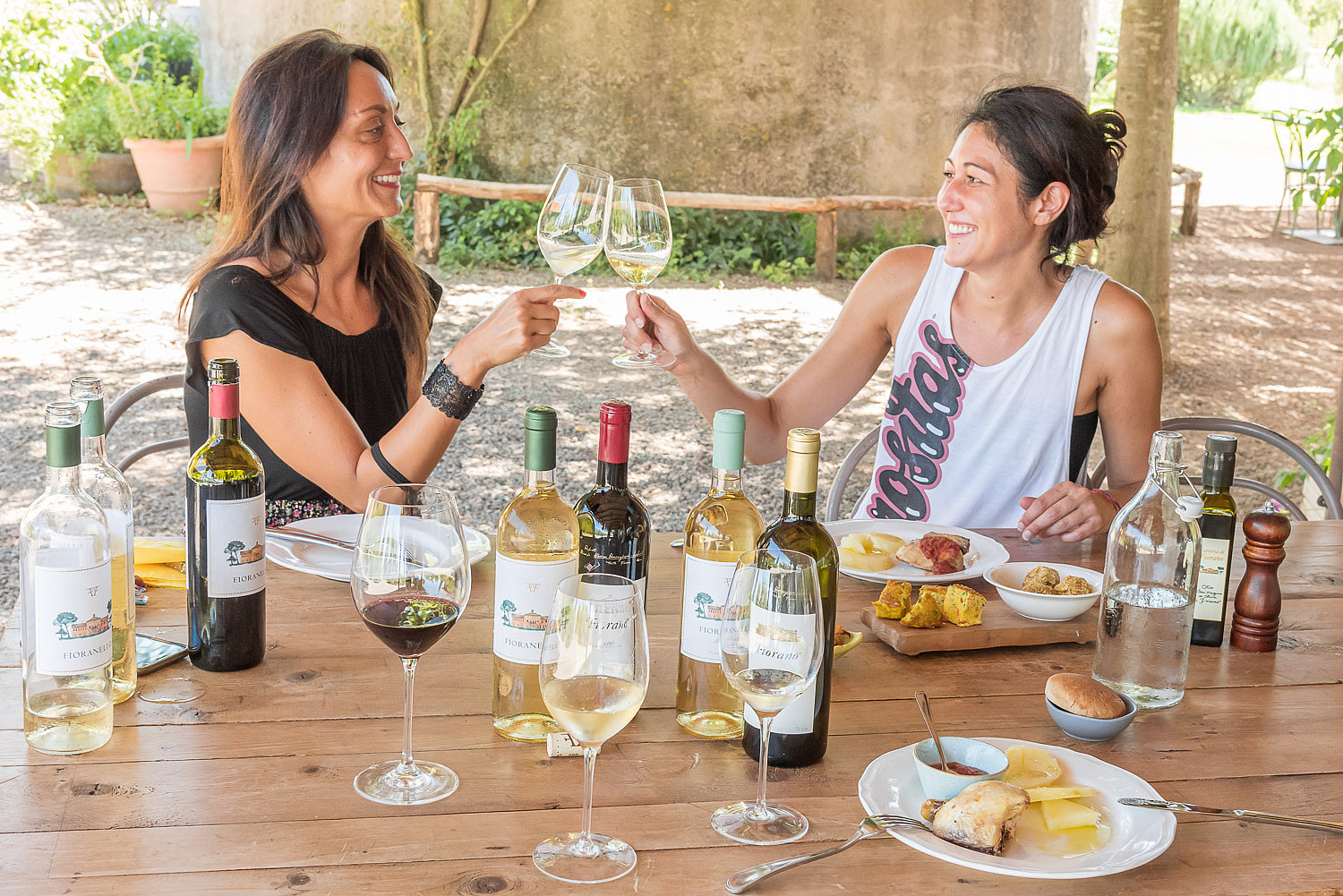 Zwei Frauen stoßen mit ihren Weingläsern an, mehrere Flaschen Wein stehen auf einem Tisch