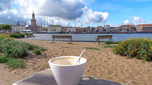 Kaffeetasse auf Holzbank an einem Flussstrand mit Skyline einer Stadt im Hintergrund
