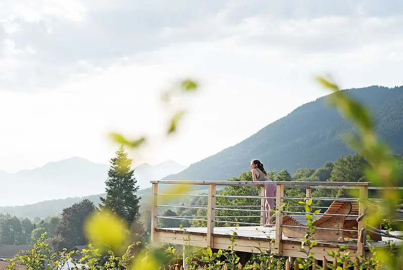 Eine Frau lehnt an der Brüstung einer Holzterrasse vor einem idyllischen Bergpanorama