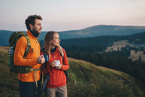 Man sieht einen Mann in gelber Jacke und eine Frau in roter Jacke mit Rucksäcken auf dem Rücken und Tassen in der Hand. Es ist Dämmerung, man sieht im Hintergrund die Landschaft des Schwarzwalds.