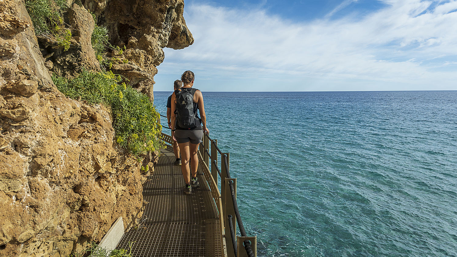 Zwei Personen laufen einen Weg direkt an einer Felswand über dem Meer entlang