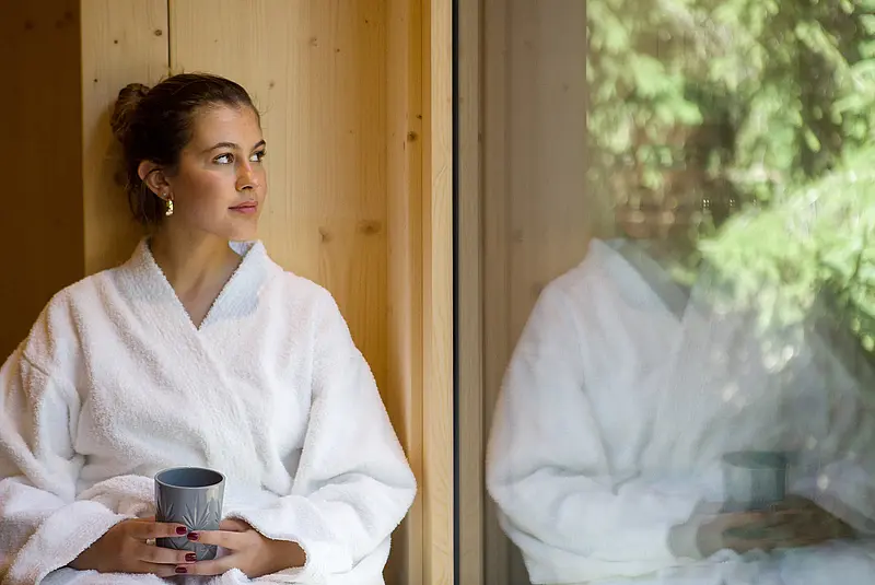 Eine mit einem weißen Bademantel bekleidete Frau sitzt mit einer Tasse in der Hand vor einer Fensterfront mit Blick ins Grüne
