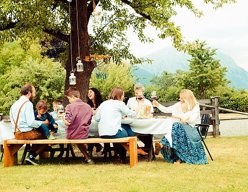 Mehrere Erwachsene und mehrere Kinder sitzen um einen großen Tisch in einem Garten. Sie essen und trinken