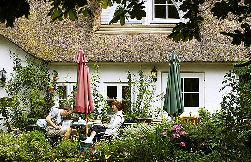 Zwei Menschen sitzen an einem Teetisch vor einem Haus mit Reetdach