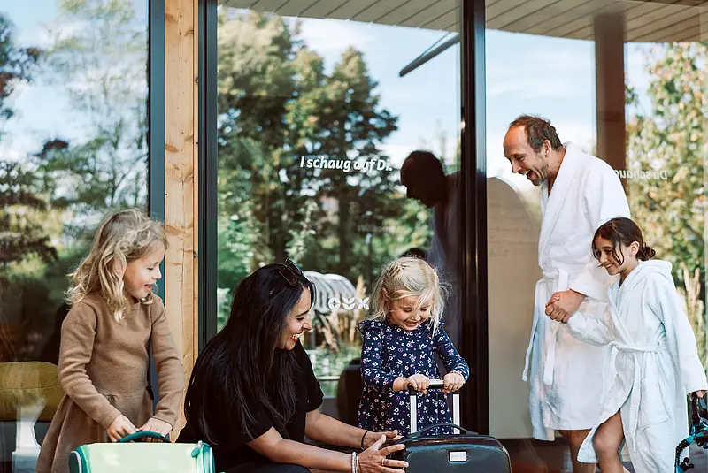 zwei Frauen und ein Kind sitzen mit Koffern vor einem Hoteleingang und sprechen mit einem Vater und seiner kleinen Tochter (beide mit weißen Bademänteln bekleidet)