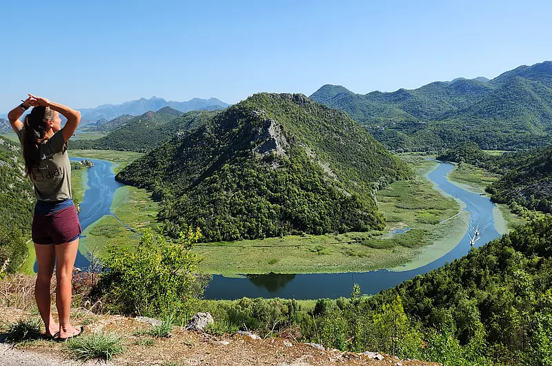 Eine Frau in Wanderklamotten steht auf einer Anhöhe und blickt auf eine Flussschleife, die sich um einen Berg windet