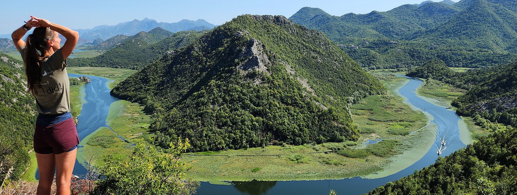 Eine Frau in Wanderklamotten steht auf einer Anhöhe und blickt auf eine Flussschleife, die sich um einen Berg windet