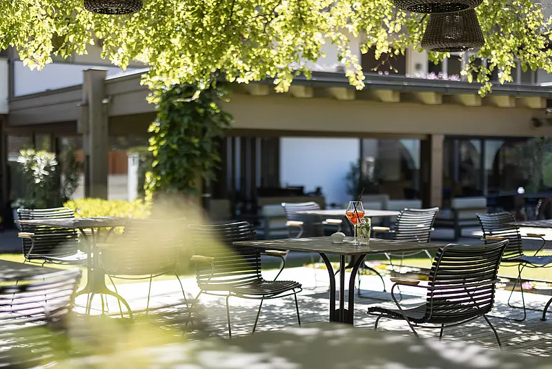 Tische und Stühle stehen auf einer Terrasse unter dem Blätterdach eines Baumes