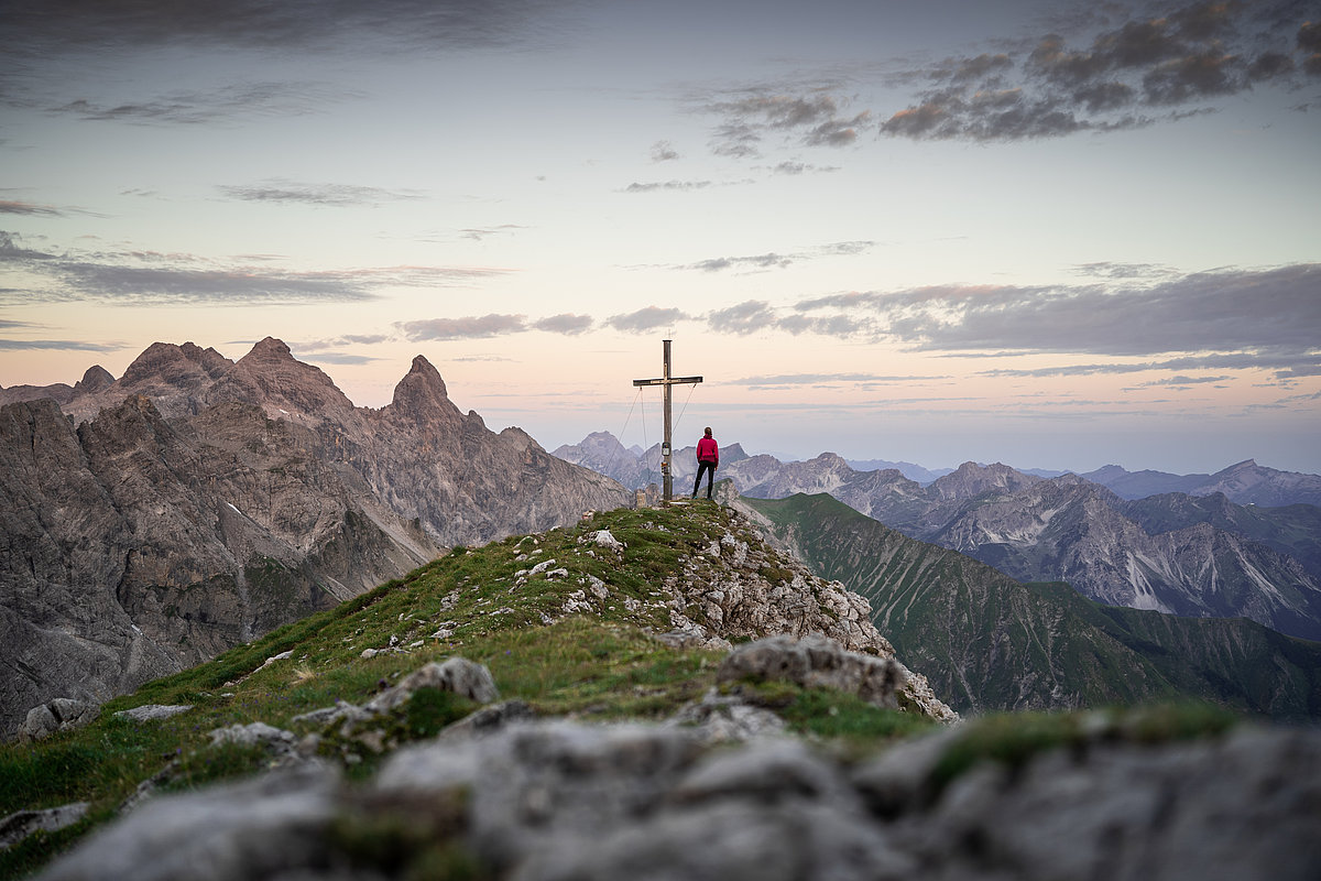 Ein Mensch steht neben einem Gipfelkreuz mit Blick auf die Alpen