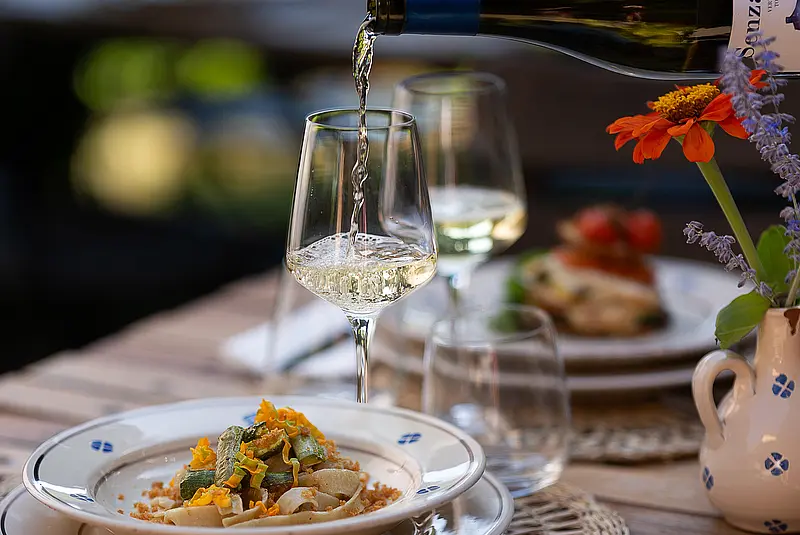 Gedeckter Tisch mit zwei mit Nudeln gefüllten Tellern und zwei halb gefüllten Weingläsern. Jemand gießt Wein aus einer Flasche in eins der beiden Gläser