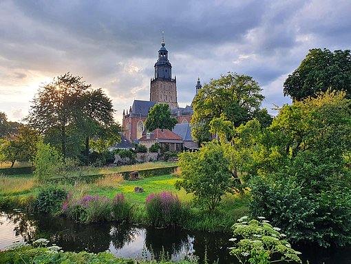 Blick über ein Flüsschen und Flussaue mit Bäumen und Wiesen auf eine Kirche