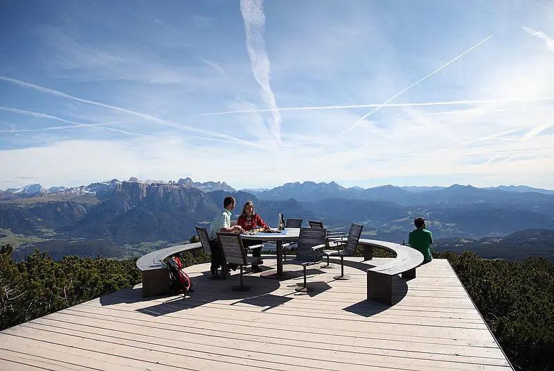 Ein Mann und eine Frau sitzen auf einer Aussichtsplattform am Tisch und essen, im Hintergrund Bergpanorama
