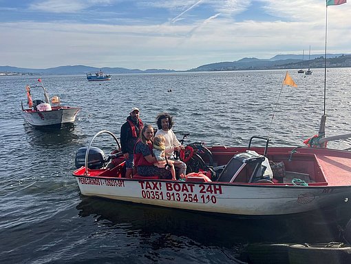 Eine Mutter und ein Vater sind mit ihrer Tochter auf einem Boot auf dem Wasser. Auf dem Boot steht: "Taxi Boot" und es steht ein weitere Mann am Ruder.