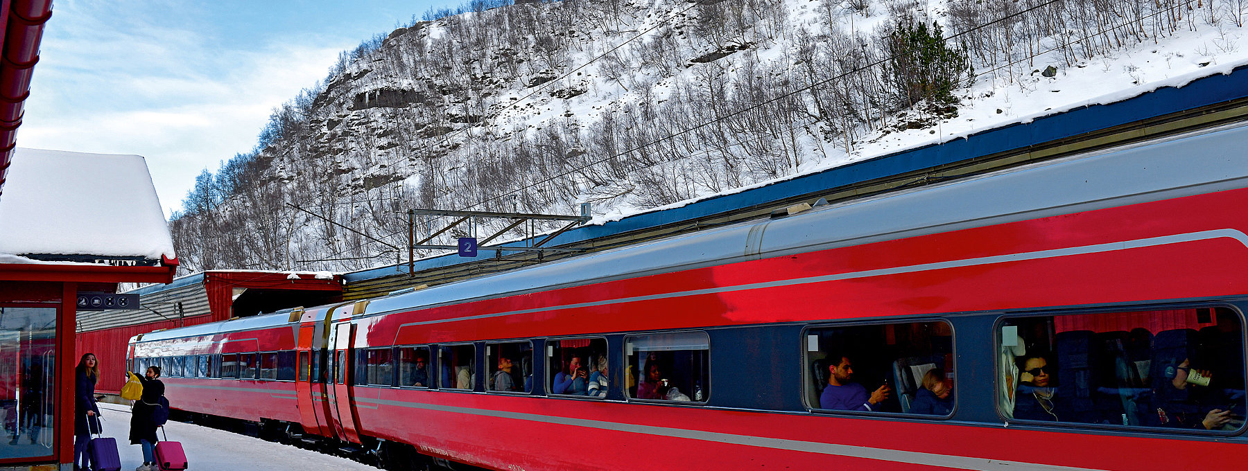 Roter Zug mit Passagieren, verschneiter Bahnsteig und Felsberg
