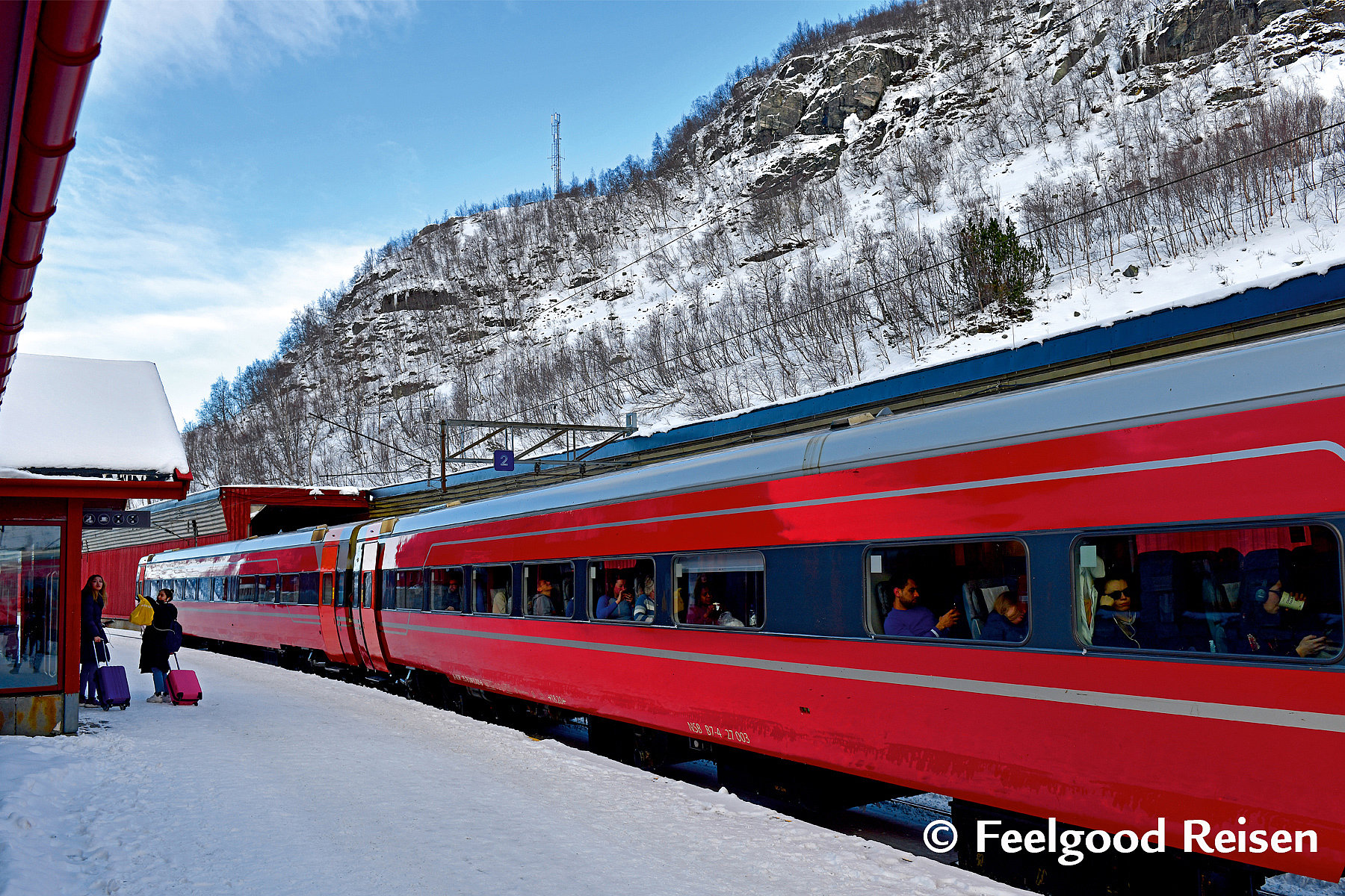 Roter Zug mit Passagieren, verschneiter Bahnsteig und Felsberg