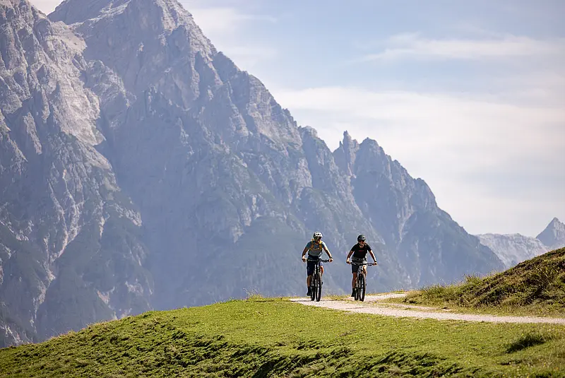 Zwei Mountainbikefahrer radeln nebeneinander auf einem flachen Weg, im Hintergund schroffe, felsige Berggipfel