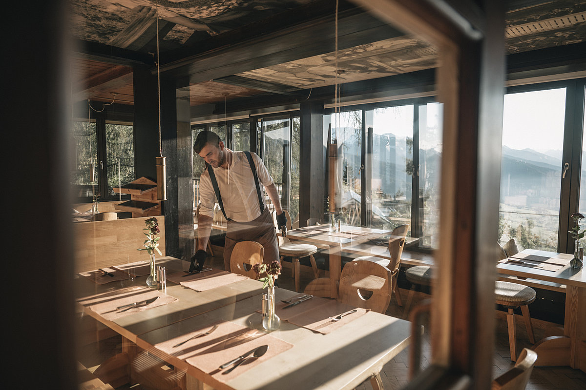 Ein Mann deck einen Holztisch in einem Speiserestaurant eines Restaurants ein. Im Hintergrund ist das Bergpanorama durch die großen Fenster erkennbar.