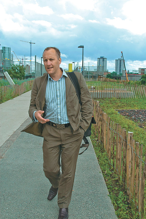 Ein Mann im braunen Anzug läuft über einen Fußgängerweg, London im Hintergrund