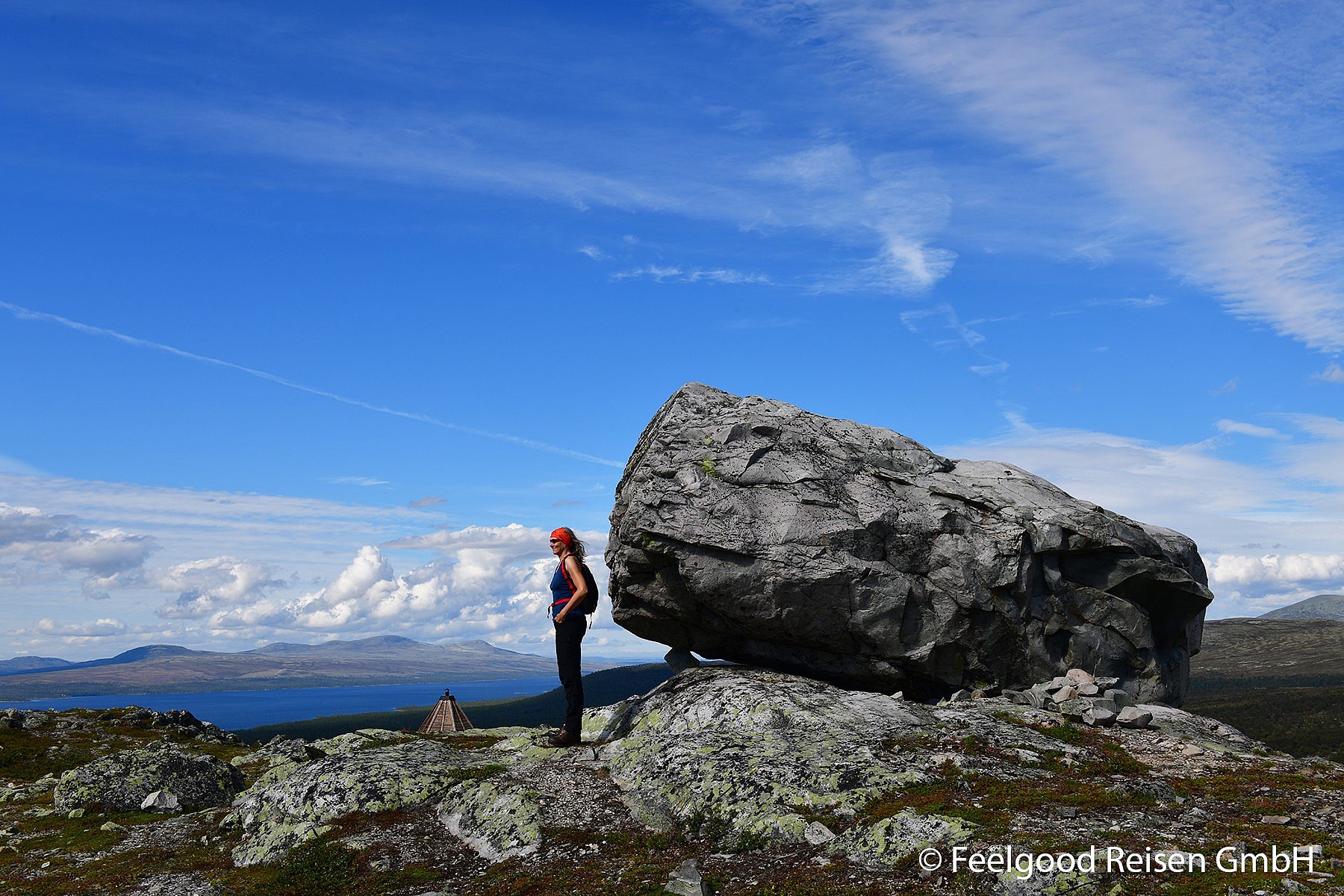 Eine Frau steht auf dem Gipfel eines Berges und betrachtet das Bergpanorama, hinter ihr befindet sich ein großer Fels